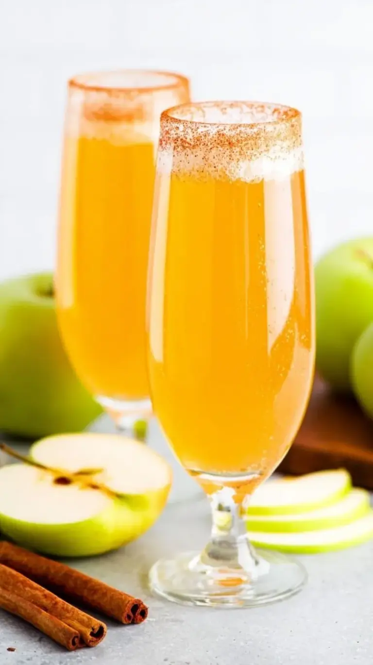 Four champagne flutes filled with golden apple cider mimosas, rimmed with cinnamon sugar and garnished with apple slices and cinnamon sticks.